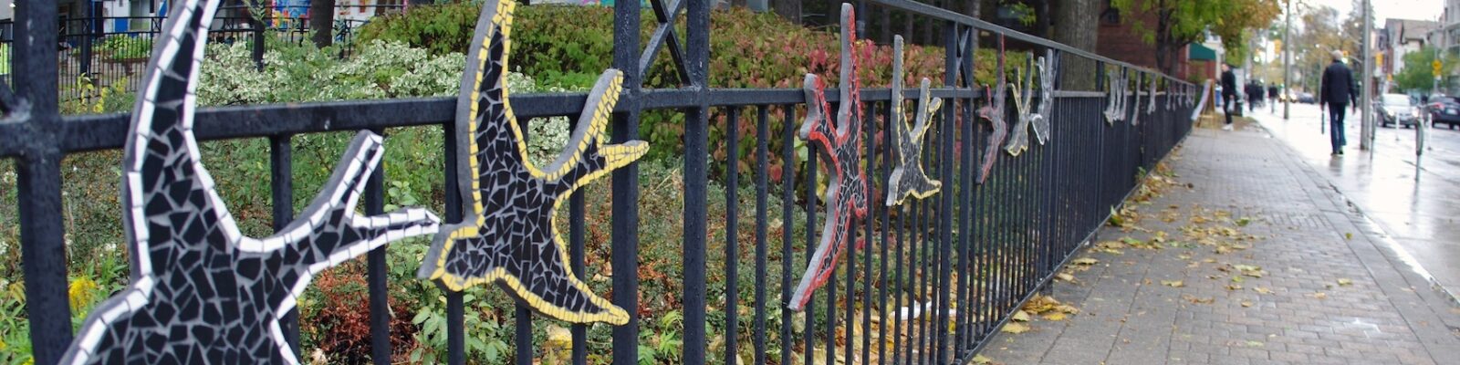 A series of ceramic and glass mosaic swallows, installed on a low black fence, recede into the distance. There are shrubs and a building in the background. On the right side of the photo, a sidewalk runs alongside the fence, and appears to narrow into the distance.