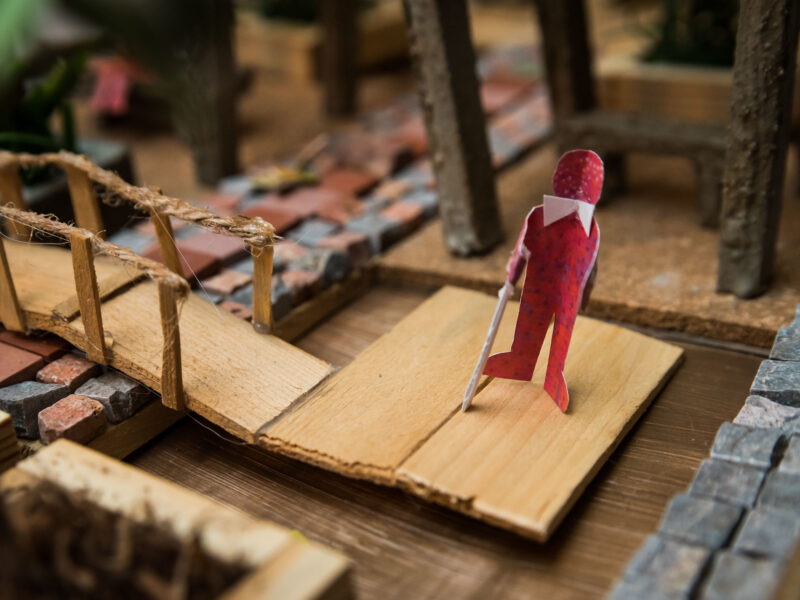 Close-up photo of a neighbourhood diorama focused on an individual with a guide cane next to a bridge.