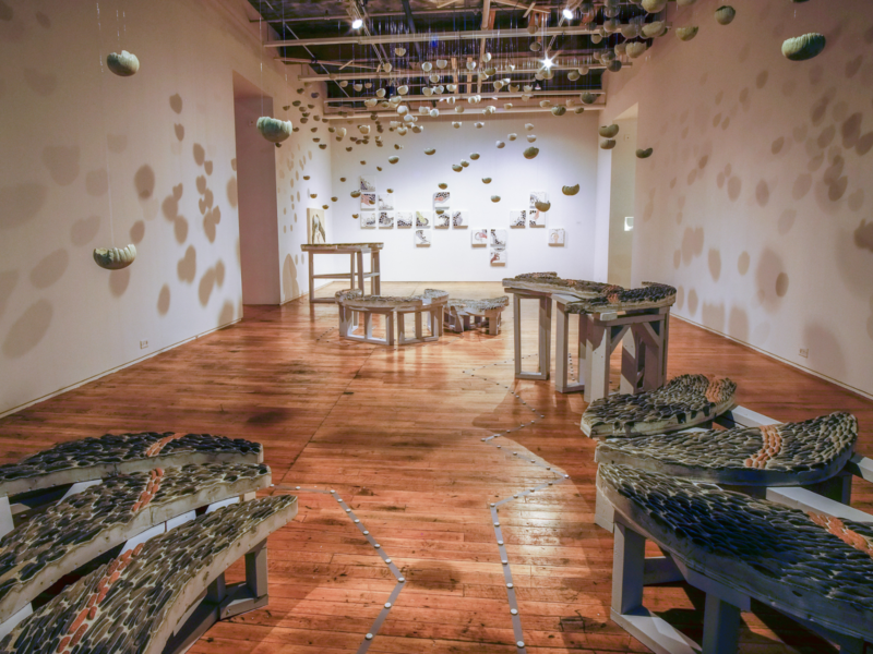 art gallery exhibit featuring multiple pebble mosaic benches of varying heights, multiple glass and ceramic mosaics, and multiple small suspended vessels that cast shadows onto the gallery walls.