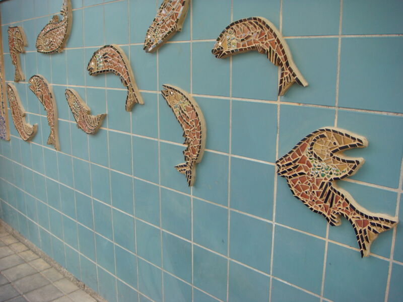 Multiple small mosaics in the shape of salmon decorate a blue tiled wall.