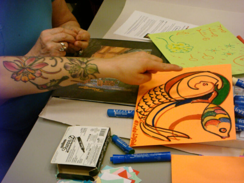 A hand pointing at a drawing on a table. Drawing depicts a merperson and a fish.