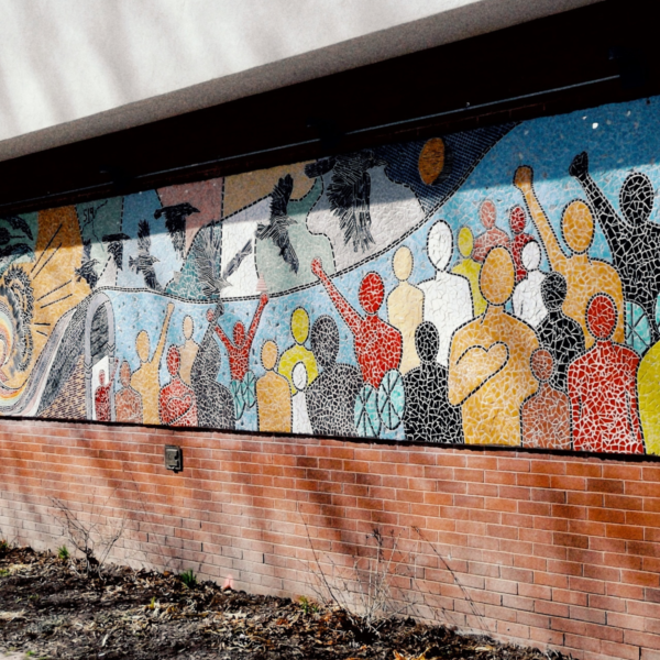A long mosaic mural installed on the side of a building depicts silhouettes of people, birds flying overhead in the sky and a large tree and rainbow.