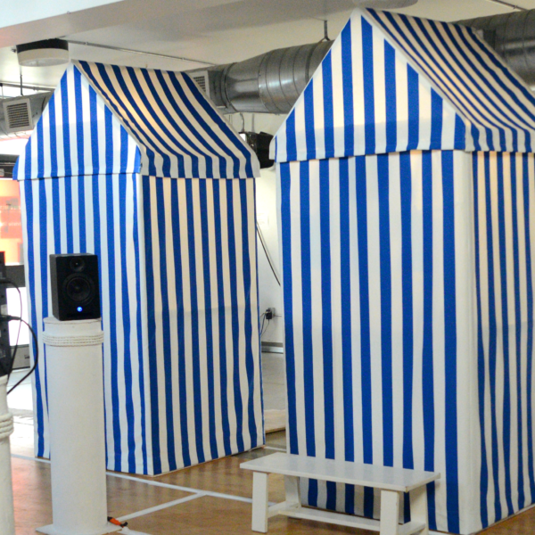 A performance space set up with three small changing huts surrounded by tall columns topped with audio speakers.