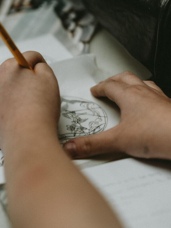 Close-up of a person’s hands, drawing dragonflies and waves on tracing paper. Beneath the tracing paper is a printed sheet of many different mosaic design templates.