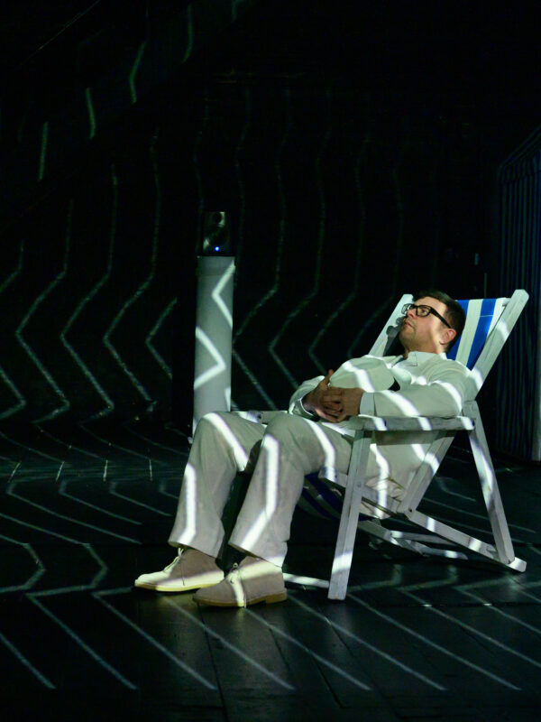 Tristan sits in a beach chair on a dark stage. A white light projection of zig-zagging lines are projected onto him.