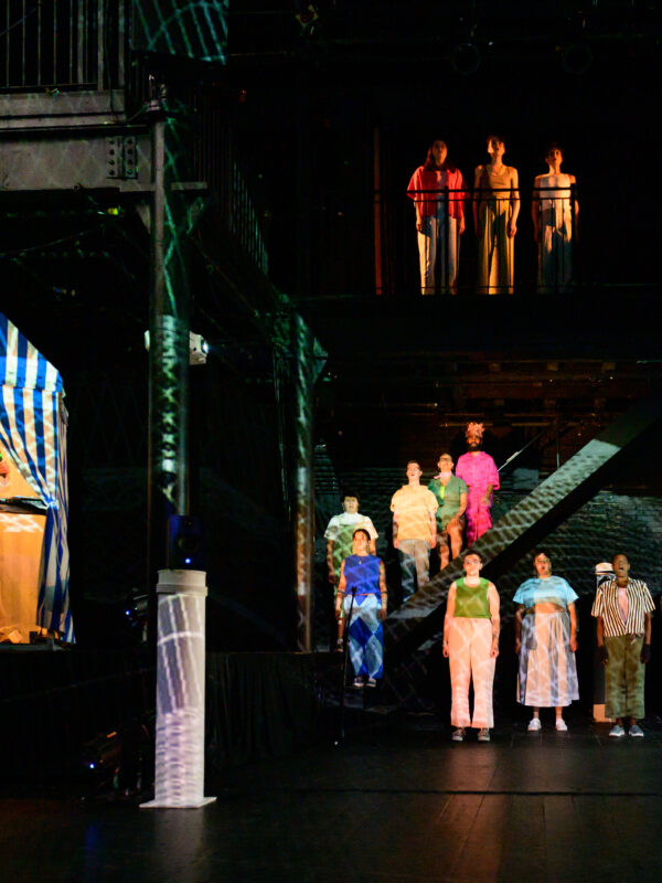 The Epic Choir of Traceland stands in formation on the stage, stairs, and upper balcony, facing the audience. Tristan stands at his computer in the beach hut, and white lines are projected over the stage and performers.