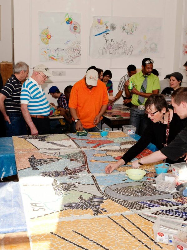 Many people in a crowded room work and socialize around mosaic panels placed on tables.