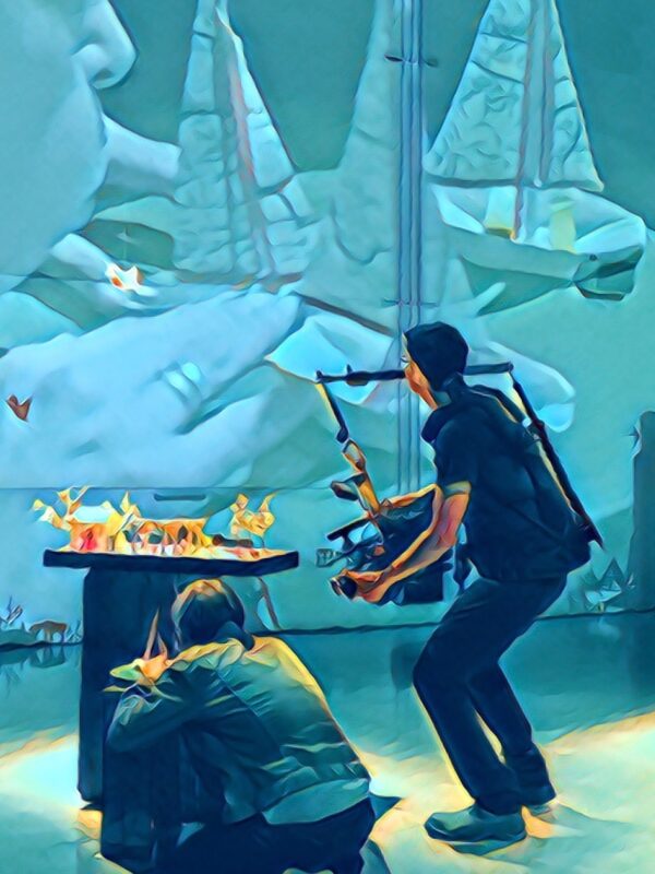 A cameraperson points their camera at an artist on the ground beside them. On the wall in front of them both, live camera footage is projected showing a close up of the artist’s hands, touching tiny paper sailboats. She blows on the sailboats. The image has been treated with a filter, giving it a smooth, brush stroke-like texture and blue glow, creating an “underwater” feel to the image.