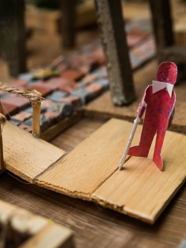 Close-up photo of a tiny hand crafted outdoor environment. A paper cut out person wearing a bow tie and holding a white cane stands at the end of a pedestrian bridge. 