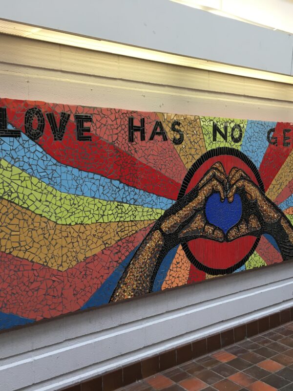 A large colourful mosaic depicting hands formed into a heart shape, with text that reads, “love has no gender”.