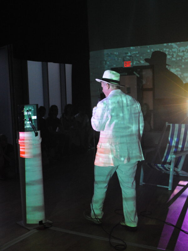 Tristan R. Whiston, in performance, walking through a performance space lit solely by projected video of an ocean.