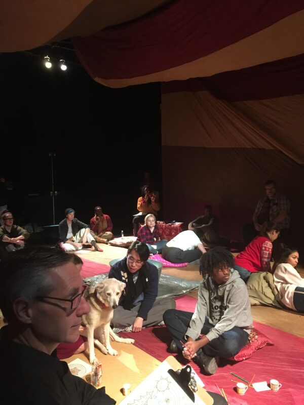 A room filled with people, and a dog, sitting on pillows and blankets on the floor.