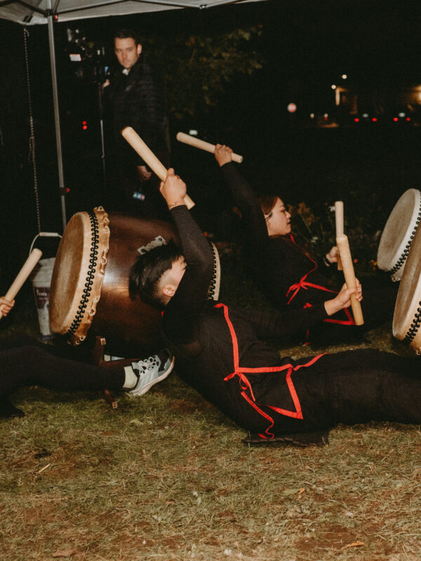 Three taiko drummers sit on the ground playing large barrel shaped drums. All three are leaning back in unison, about to swing and hit the drums.