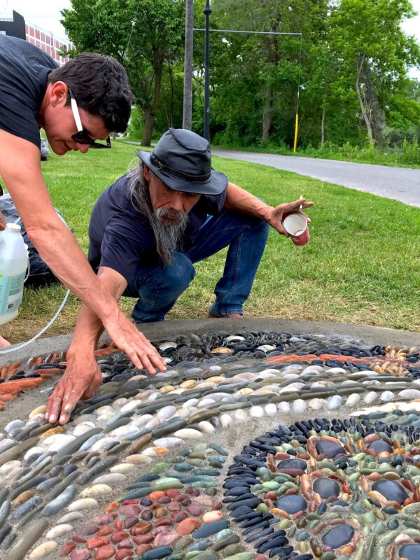 Photo of two artists kneeling down to touch and observe a pebble mosaic. One holds a jug of liquid and the other holds a cigarette and empty coffee cup. 