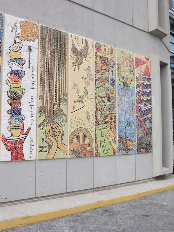 A large mosaic mural on an exterior concrete wall is made up of seven panels. Some text reads, “Support, connection, balance. Access 4 all. All kinds of families.”