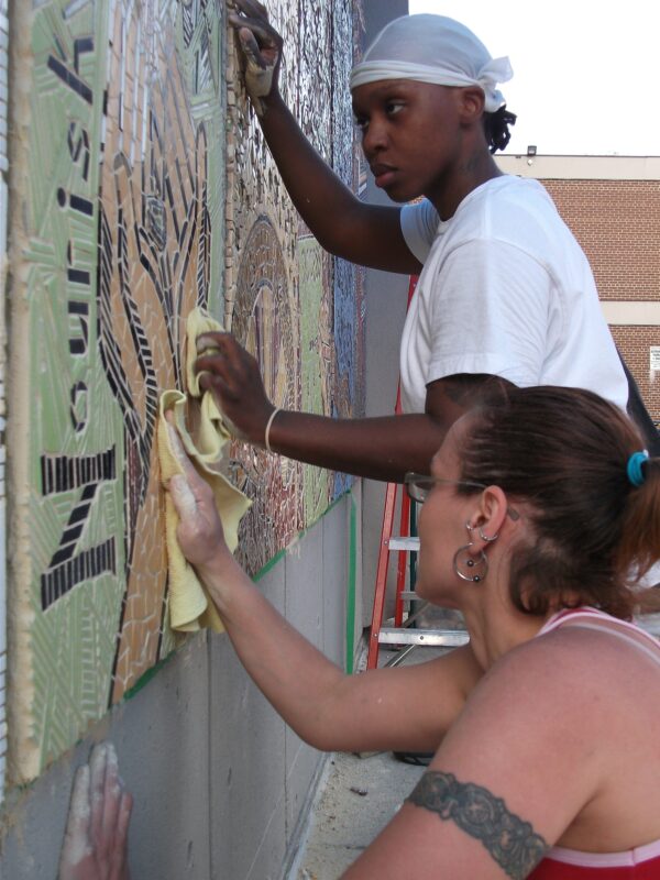 Two people use rags to polish an outdoor mosaic mural.