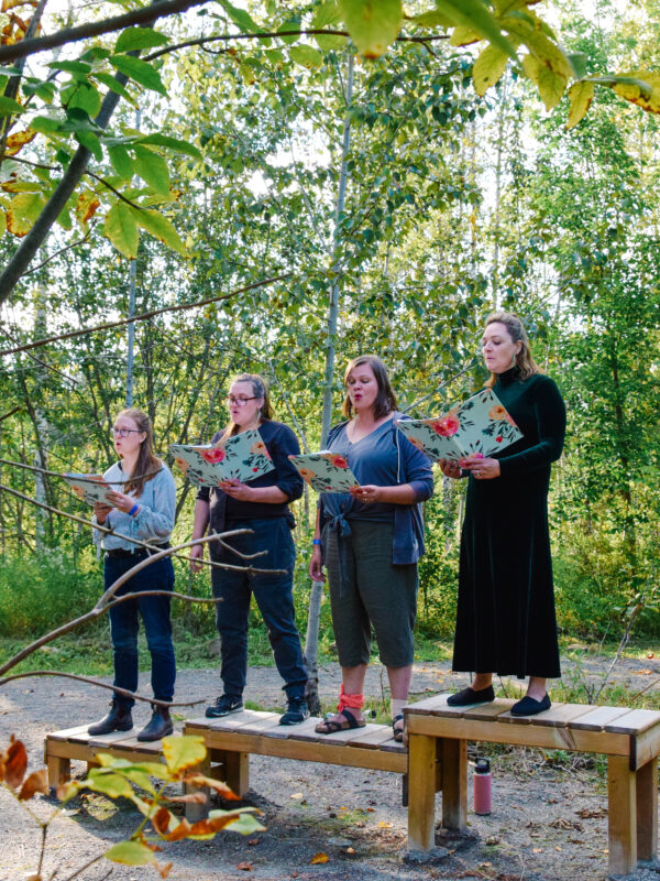 Four singers perform in the forest on a staggered wooden podium.