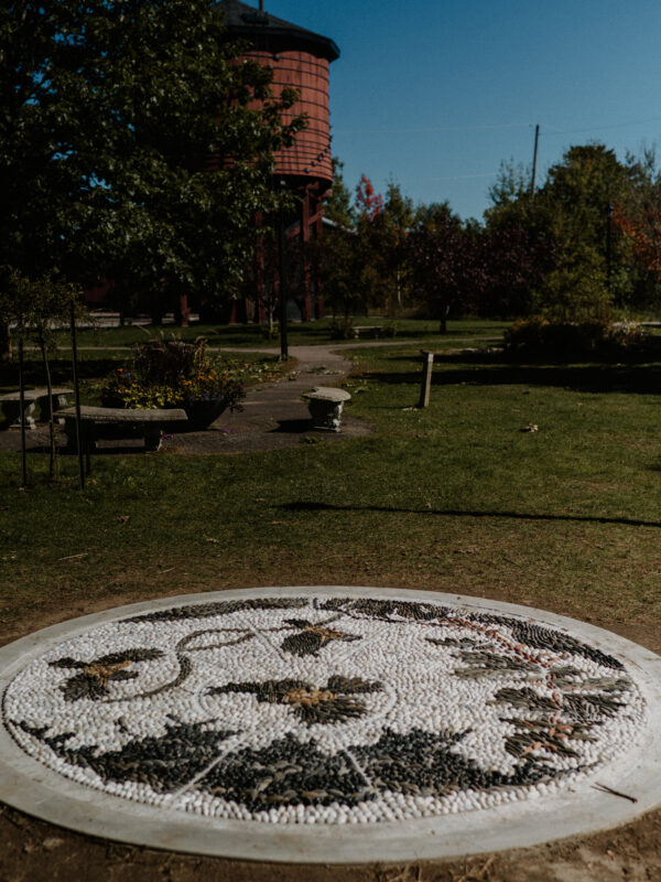 A large pebble mosaic in a park. The mosaic is surrounded by greenery, park benches, and an old wooden water tower is viewable in the background.