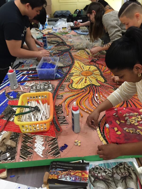 Multiple people work on a large mosaic at a table covered in art supplies.