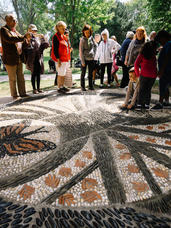 People stand on and around a large pebble mosaic that depicts a monarch butterfly and a bumblebee.
