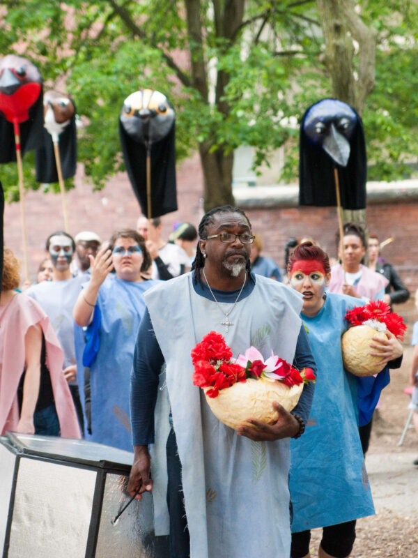 A procession of performers wearing blue and pink frocks hold large bird puppets, flowers, and a prop coffin.