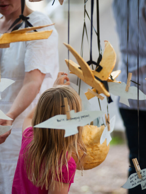A child engages with an art installation of 3D paper mâché birds and 2D paper with handwritten messages on them, suspended mid-air with black ribbon.