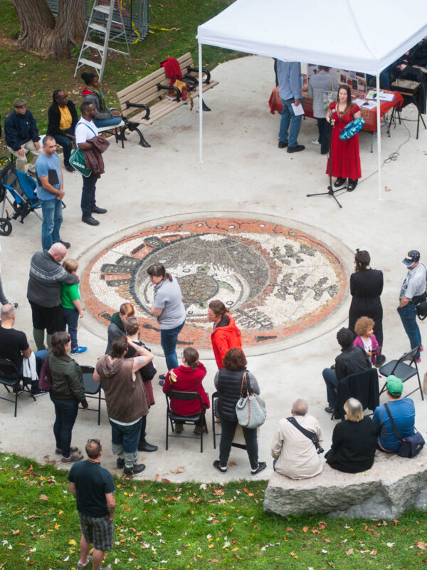 A performer sings and drums, surrounded by a crowd gathered around a large pebble mosaic.