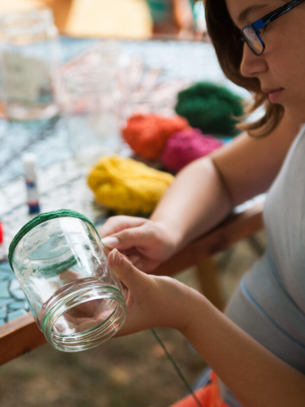 A child decorates a glass jar with yarn.