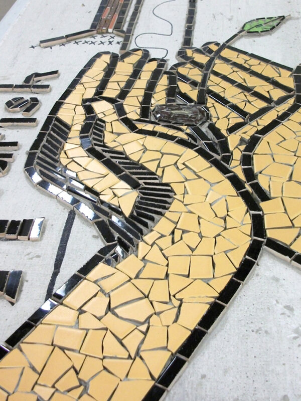 An in-progress mosaic sits on a table. The mosaic depicts a set of hands cupped together, holding a plant seedling, next to the word “Nourish”.