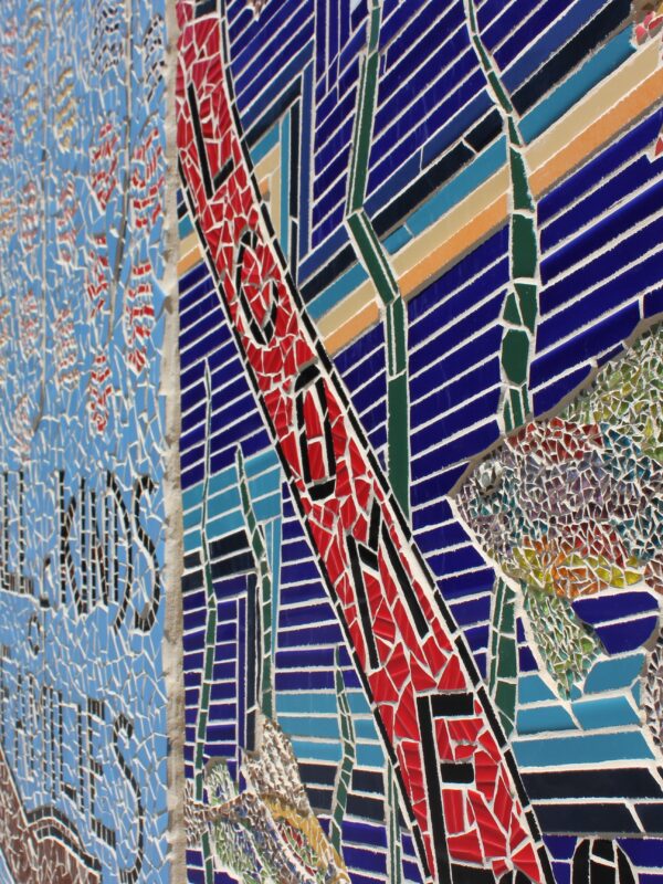 Close-up of a large, outdoor mosaic mural. Most visible section in this shot features rainbow coloured fish.