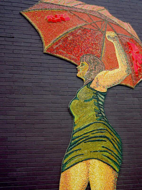 Photo of a mosaic of a woman with her fist in the air holding a red umbrella on a brick wall.