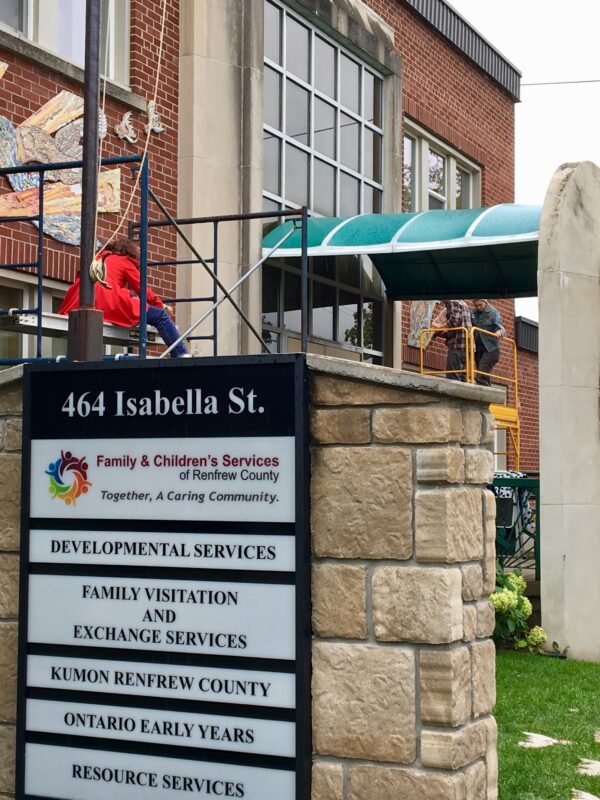 Three people on scaffolding install large mosaics on the exterior brick wall of a building. Some text on a sign next to the building reads, “464 Isabella Street. Family & Children Services of Renfrew County”.