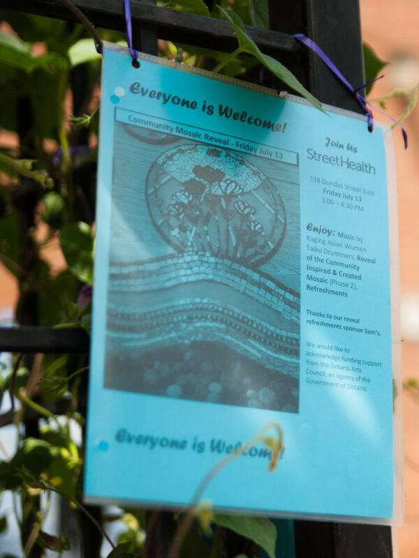 Event poster on a fence, includes image of a mosaic depicting blossoming flowers under the sun and event information. Title reads, “Everyone is welcome! Join us. Street Health”.