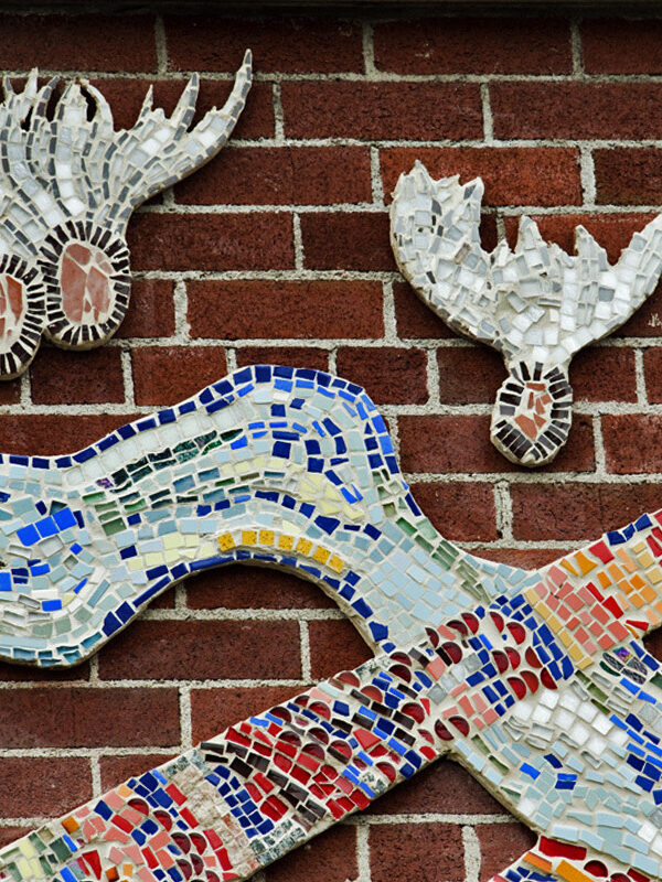 Close-up of a large colourful mosaic installed on an exterior brick wall, depicting abstract shapes and floating milkweed pods.