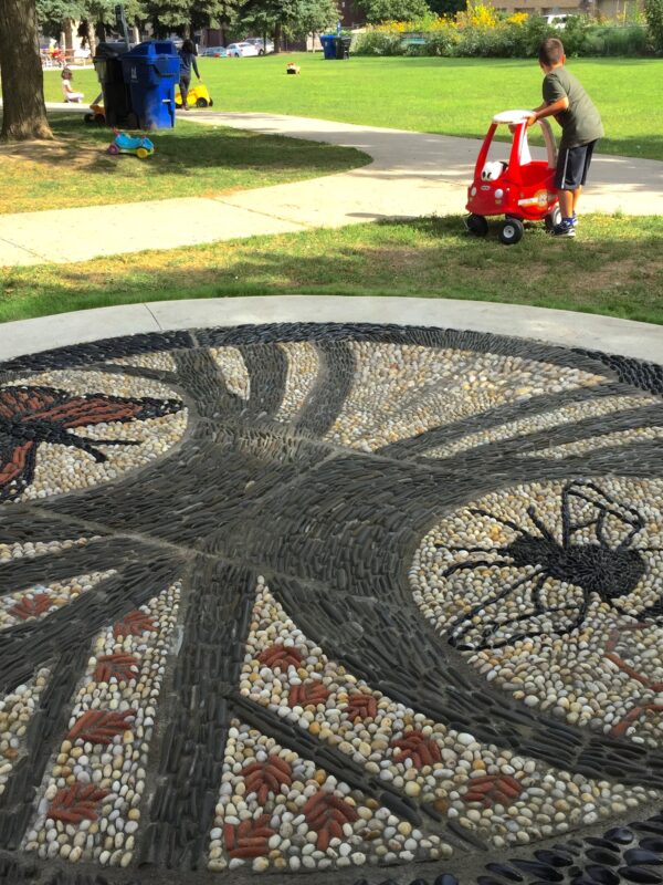 A large pebble mosaic located in a park next to a walking path. The mosaic design depicts a monarch butterfly and a bumblebee.